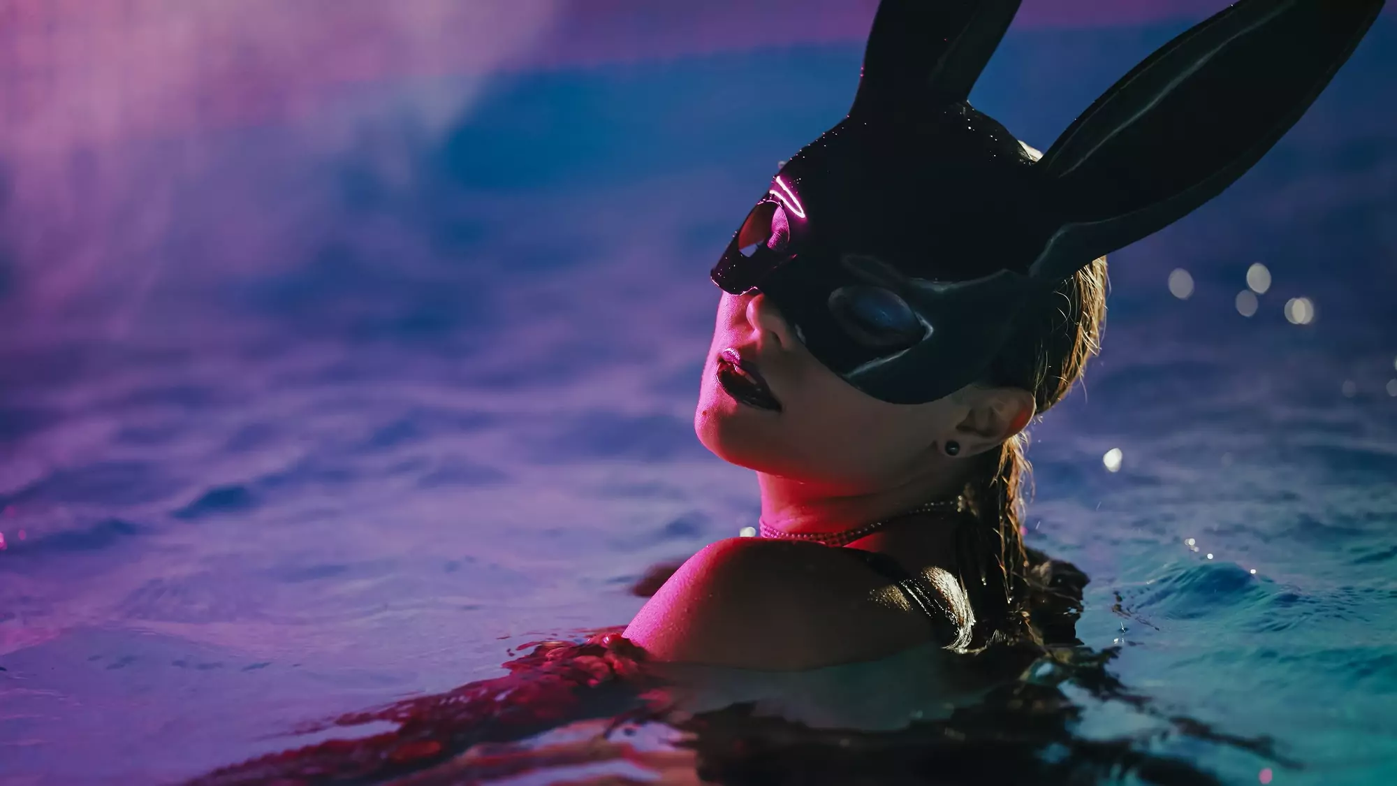 Sensual woman in leather BDSM mask moving seductively in swimming pool water under neon color light